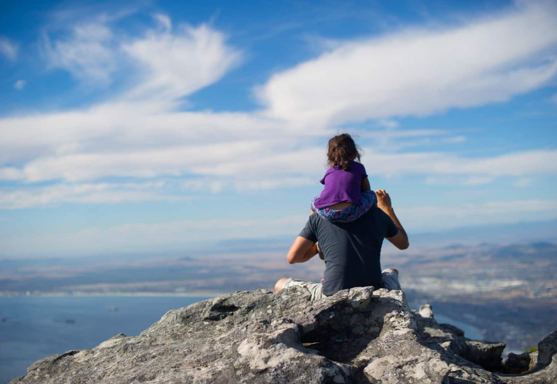 Man sitting on a rock carrying his daughter while they are both overlooking the city. The skies are bright blue and slightly cloudy.